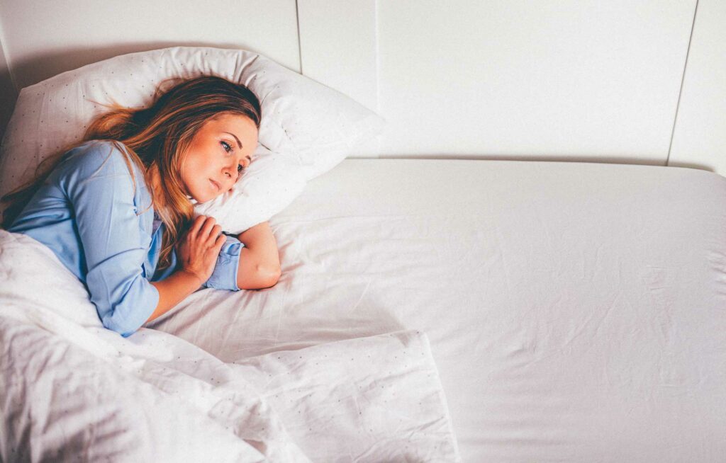 Foto einer mittelalten Frau, die alleine in einem Bett liegt und zur rechten Seite gedreht auf den leeren Platz des ehemaligen Partners blickt. Symbolisiert den Neubeginn nach Trennung und Scheidung, und die Reise zu innerer Stärke und Selbstfindung.